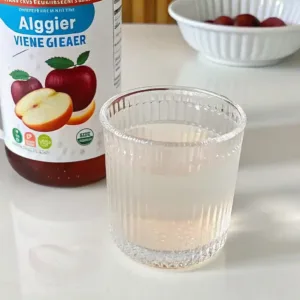 A glass of diluted pink salt and apple cider vinegar drink placed in front of a bottle labeled apple cider vinegar with the mother