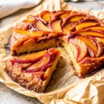 A slice of Bourbon Peach Upside Down Cake sits in front of the full cake on brown parchment paper, showing the caramelized peach topping and moist crumb.