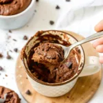 The Only Kodiak Mug Cake Recipe You'll Ever Need kodiak mug cake recipe A close-up of a fluffy and moist chocolate Kodiak mug cake in a white mug, with a hand lifting a spoonful to show the texture.