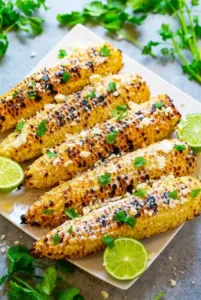 A white platter of grilled corn on the cob Mexican style, topped with crumbled cotija cheese, fresh cilantro, and served with lime wedges.