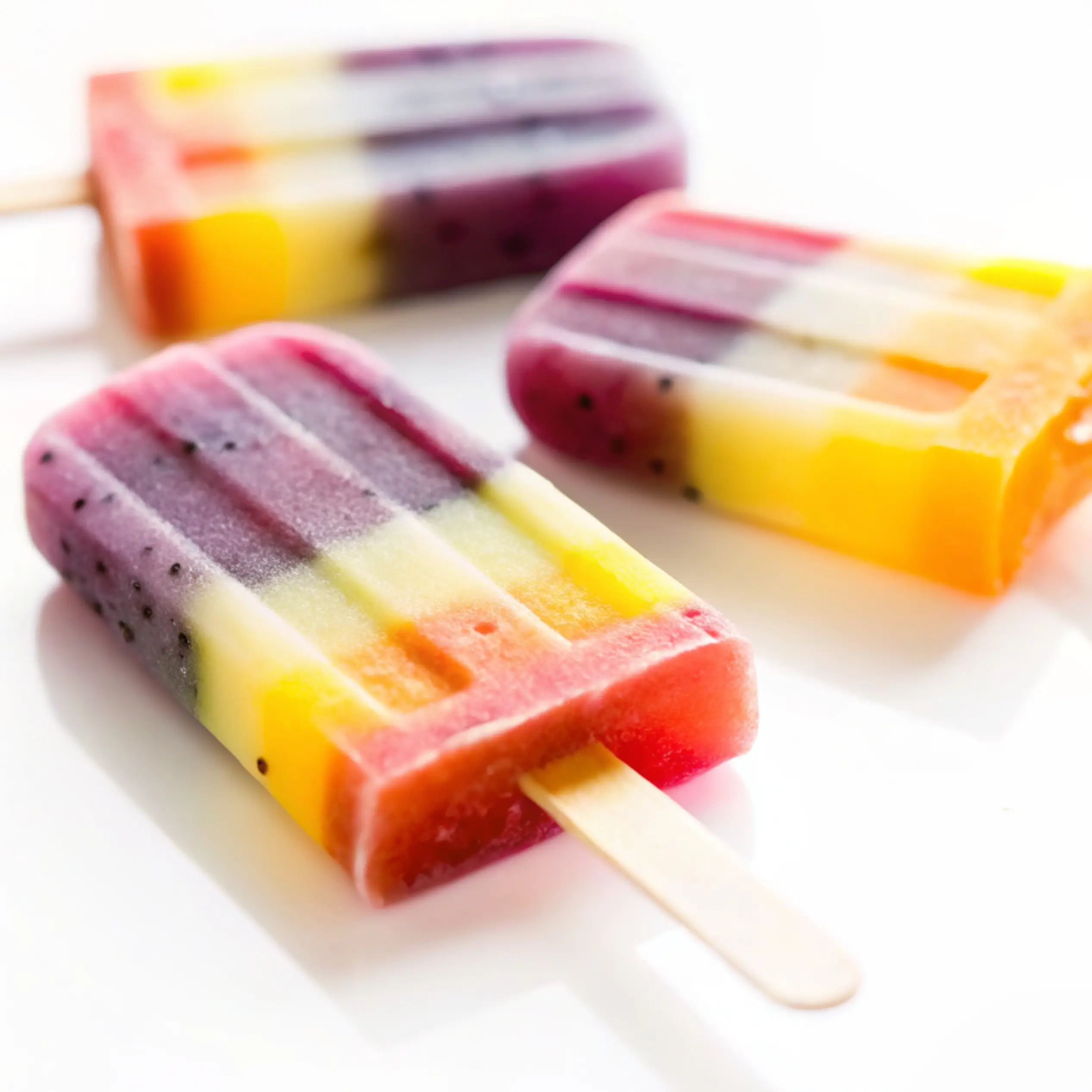 Three healthy rainbow popsicles with a vibrant dragon fruit layer, arranged on a clean white surface.