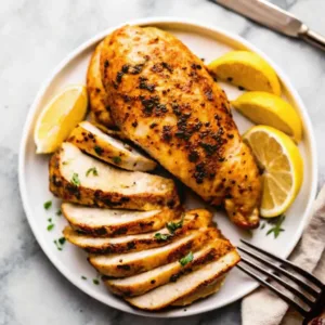 Sliced and whole juicy air fryer chicken breast on a white plate with lemon wedges and herbs