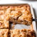 A spatula lifting a perfect slice of apple slab pie from a baking pan, showing the flaky golden crust and tender apple filling inside.