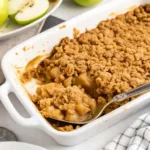Warm apple crisp baked in a white dish, topped with golden oat crumble, served with a spoon ready to scoop.