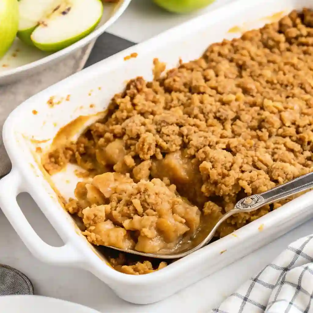 Warm apple crisp baked in a white dish, topped with golden oat crumble, served with a spoon ready to scoop.